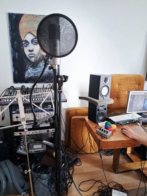 Studio corner: a microphone with a pop filter on a stand, a mixing desk and rack of outboard gear, a studio monitor on a stand, and a painting of a woman in a headwrap on the wall