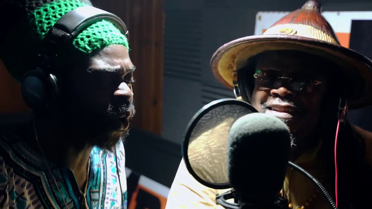 Video still: two singers in a studio, one in headphones and a green knitted hat, one in a wide-brimmed hat at the microphone