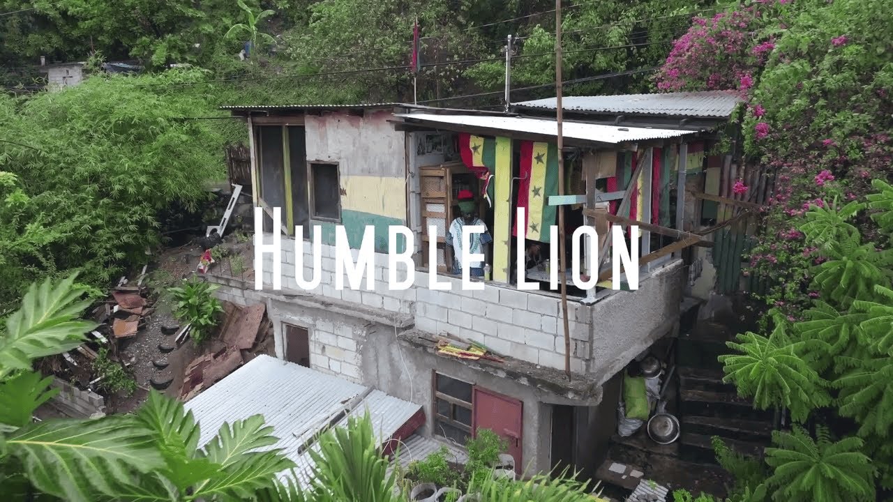 Video still: the title Humble Lion over a hillside house with red, gold and green curtains