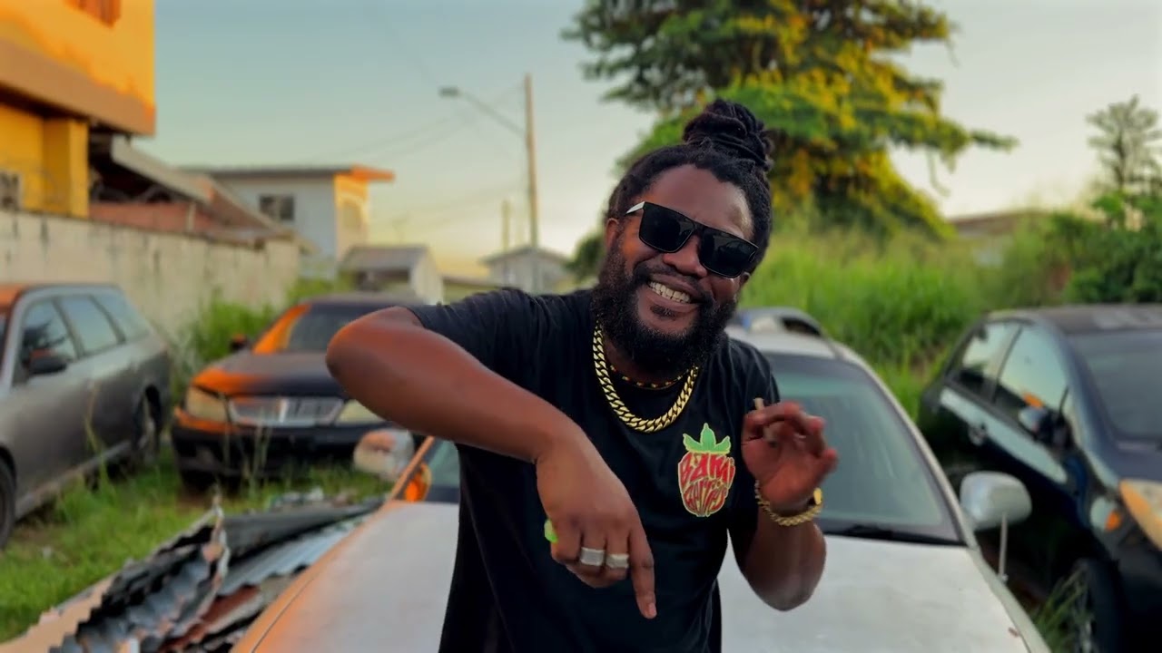 Video still: a singer in sunglasses and a gold chain in front of parked cars at sunset