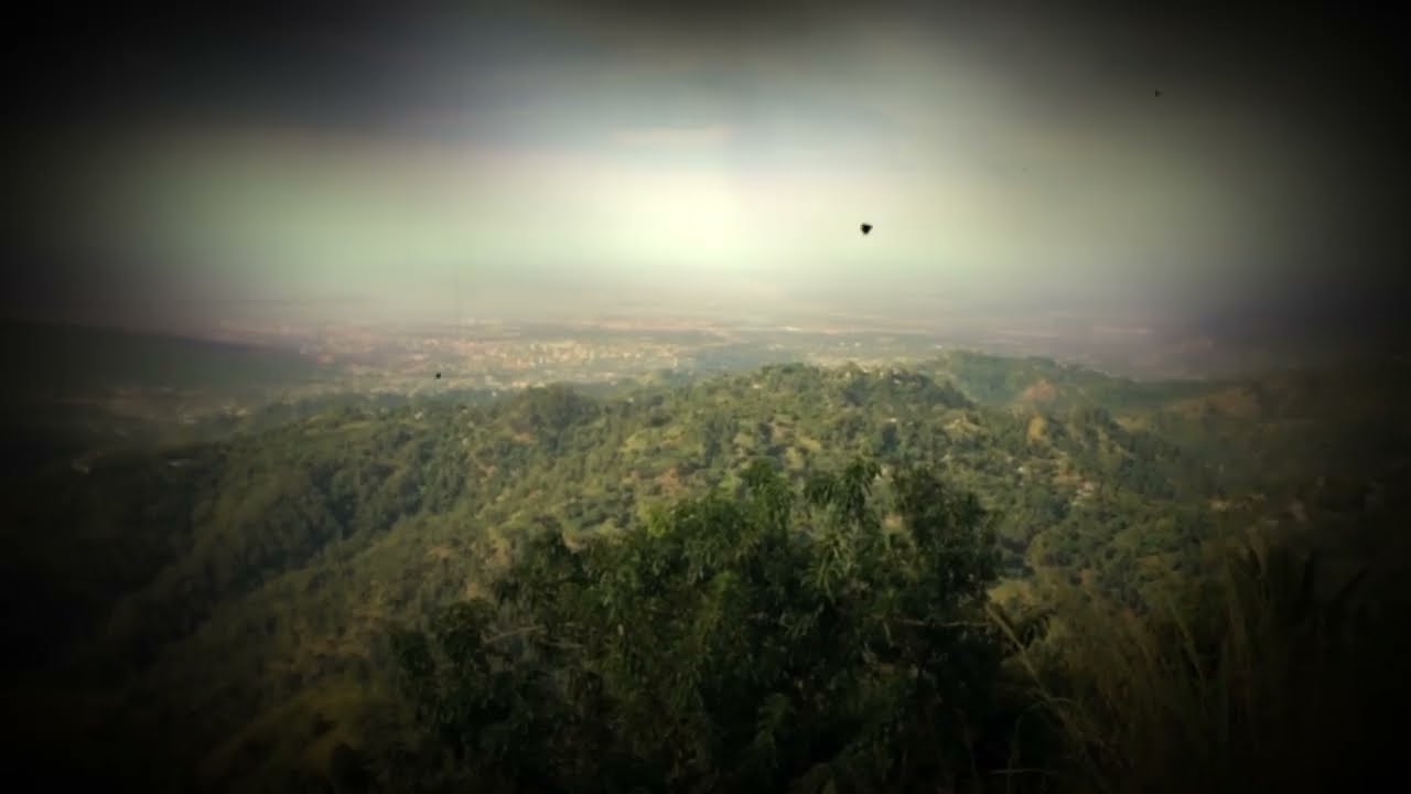 Video still: a hazy view over green hills towards a city on the plain