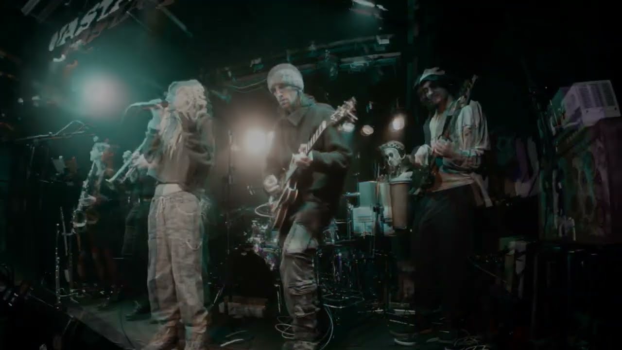 Video still: a band on a small club stage in teal light, a singer at the microphone beside a guitarist and a bassist